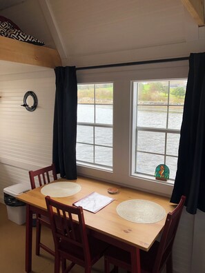 A dining area with a wooden table and chairs, two windows with a view of a body of water, and a black circular object on the wall.