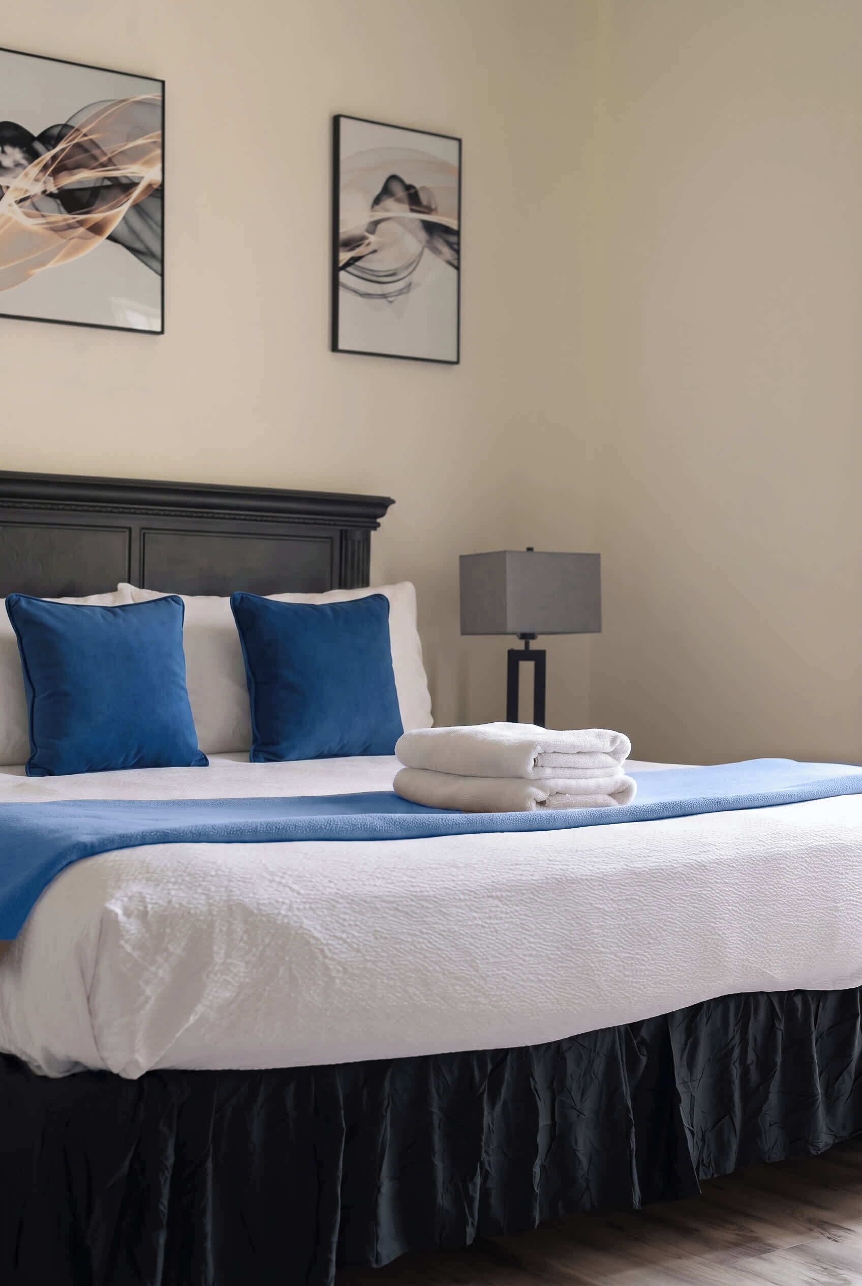 Una cama bien tendida con edredón blanco y almohadas azules, cabecera oscura, lámpara de noche y dos cuadros en la pared.