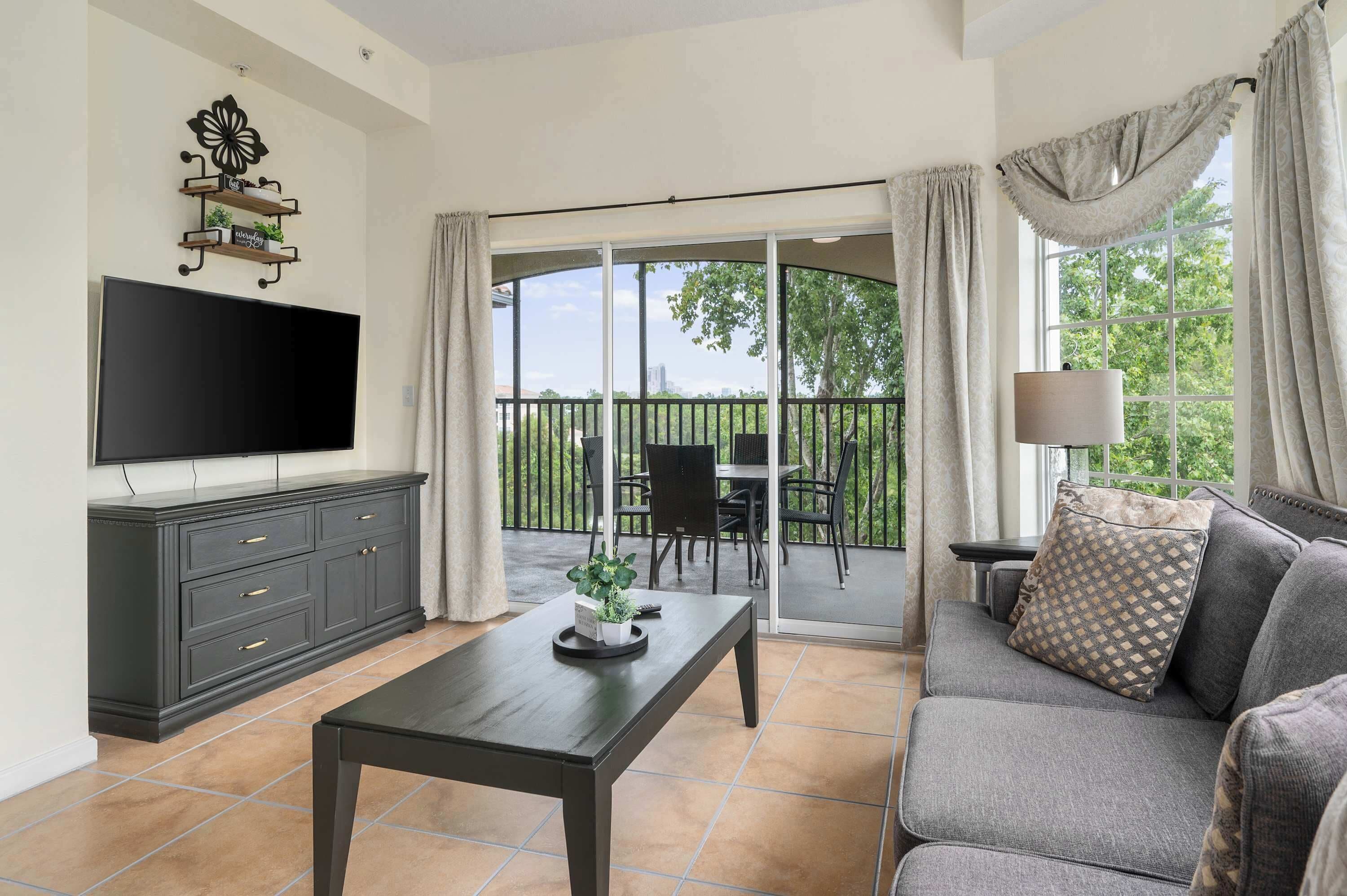 A living room with a flat-screen TV, dark wooden furniture, a sofa, a coffee table, and a balcony with a view of trees and buildings.