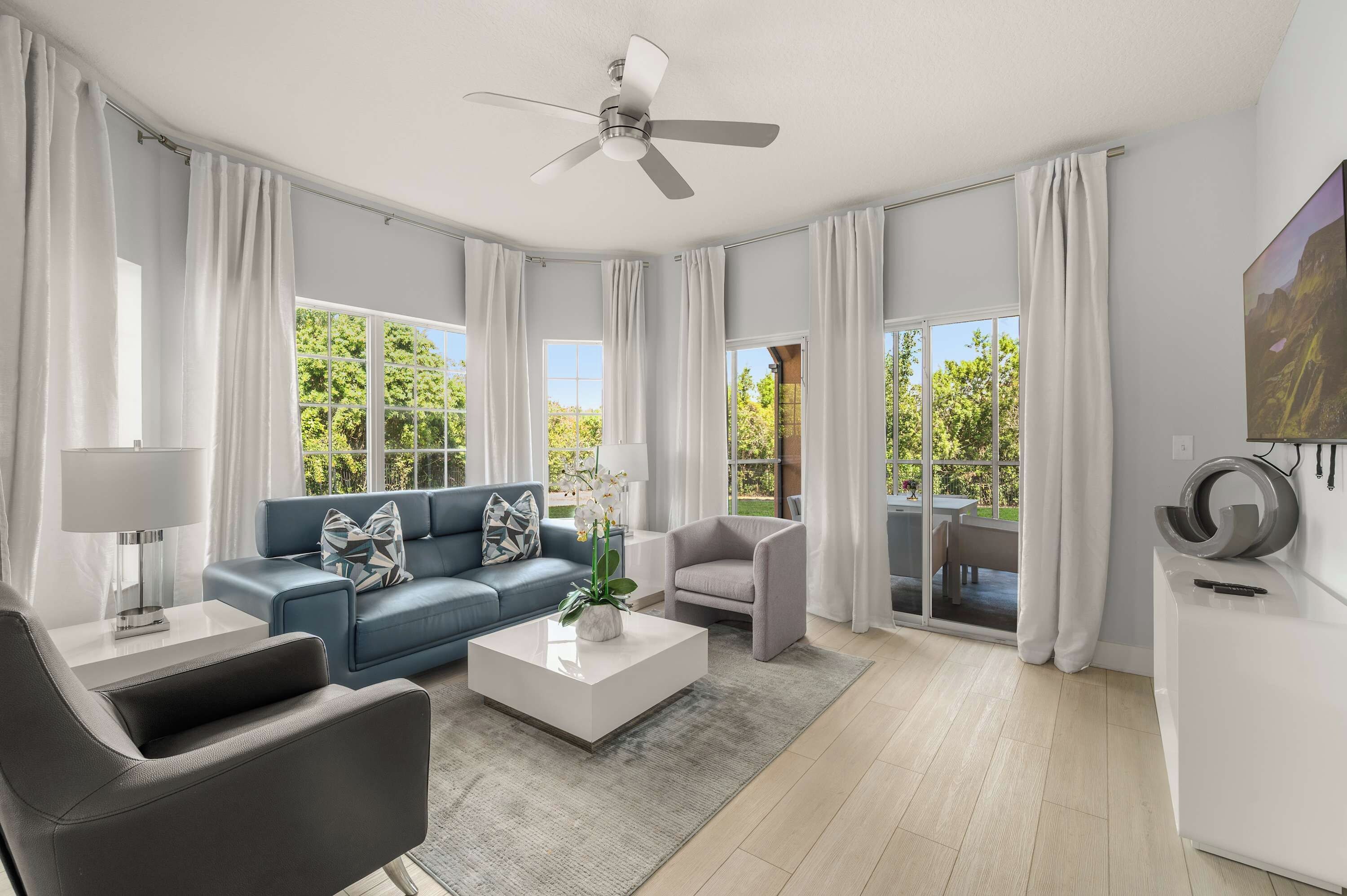 Una sala de estar moderna con un sofá azul, un sillón gris, una mesa de centro blanca, un ventilador de techo y ventanas grandes con cortinas.