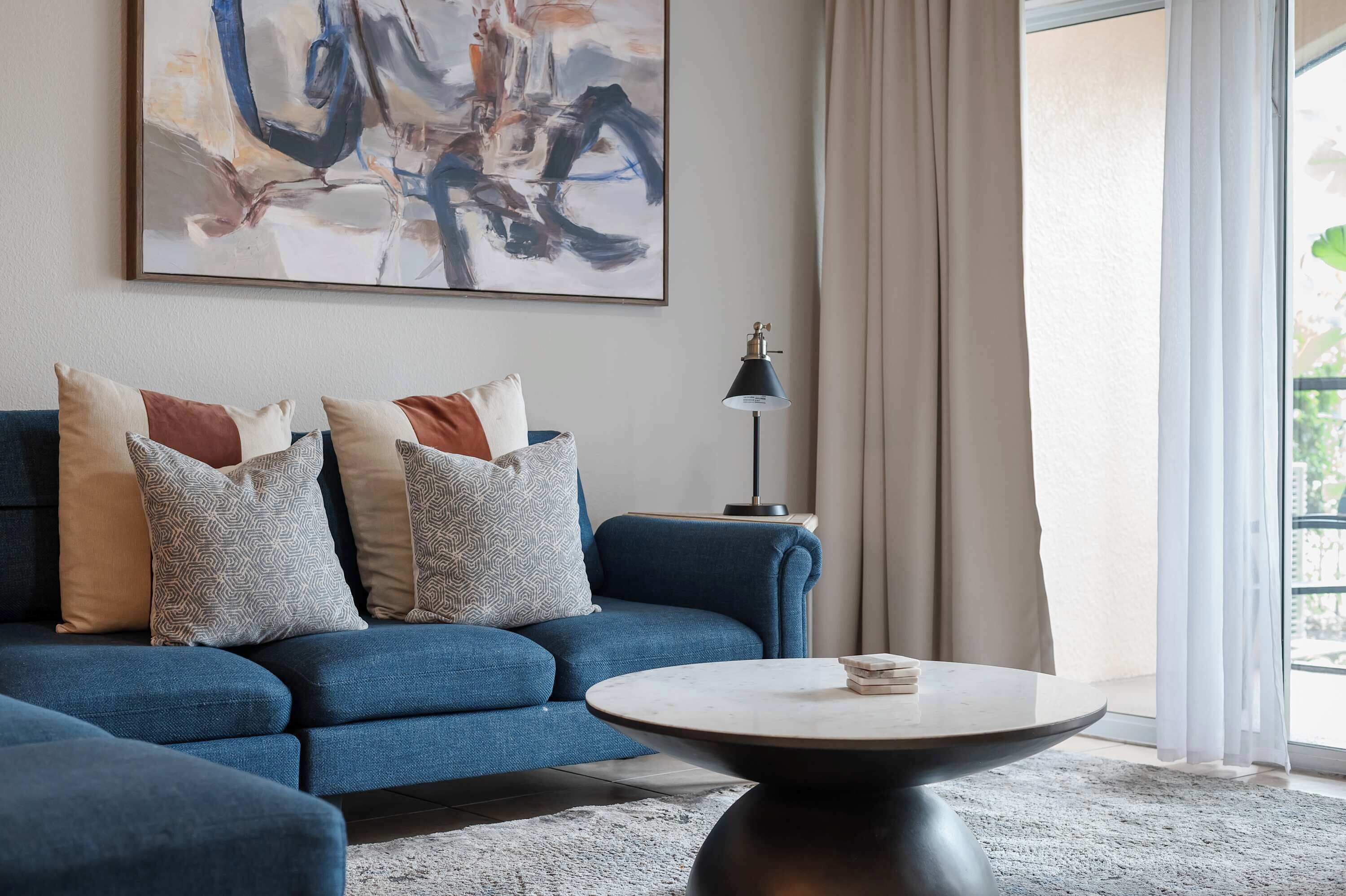 Una sala de estar con un sofá azul, cojines estampados, una mesa de centro redonda, una lámpara de pie y un cuadro abstracto grande en la pared.