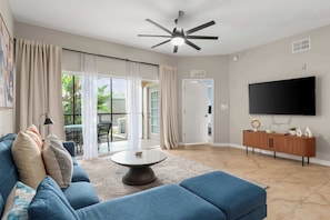 A living room with a blue sofa, a coffee table, a ceiling fan, a TV, and a sliding glass door leading to an outdoor area. - Modern Disney Escape, Near the Parks (Orlando)