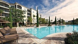 Piscina com espreguiçadeiras, um deck revestido e um edifício com varandas ao fundo.