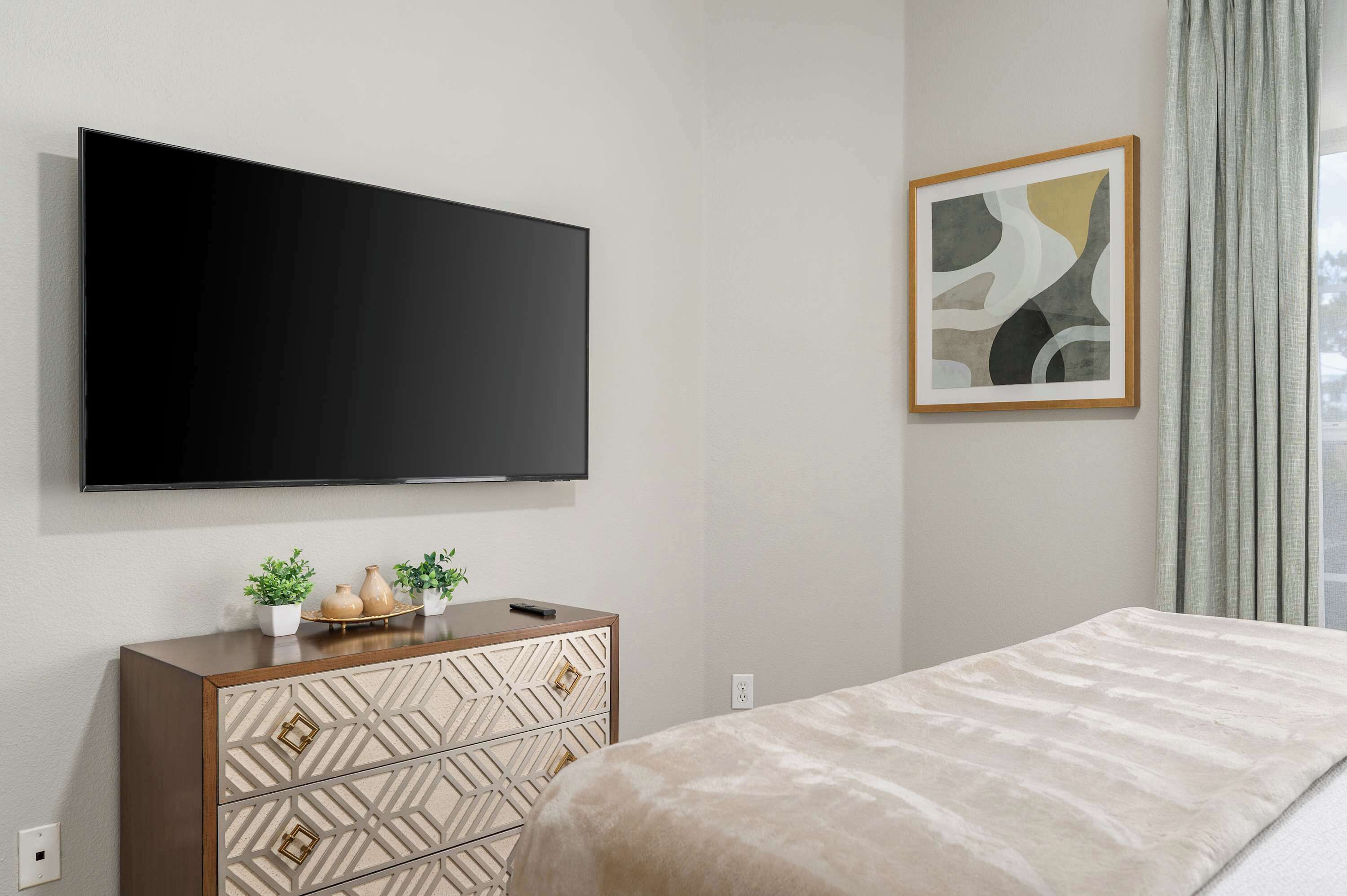 Un dormitorio moderno con una televisión de pantalla plana montada en la pared, una cama con cubrecama texturizado, un aparador de madera con paneles decorativos y un cuadro abstracto enmarcado.
