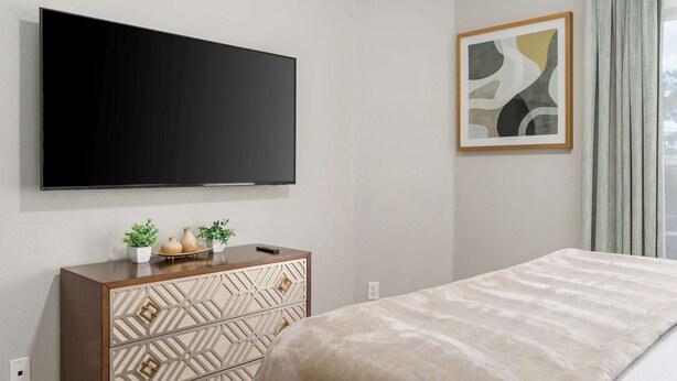 Um quarto moderno com televisão de tela plana na parede, uma cama com cobertura texturizada, uma cômoda de madeira com painéis decorativos e uma pintura abstrata emoldurada.