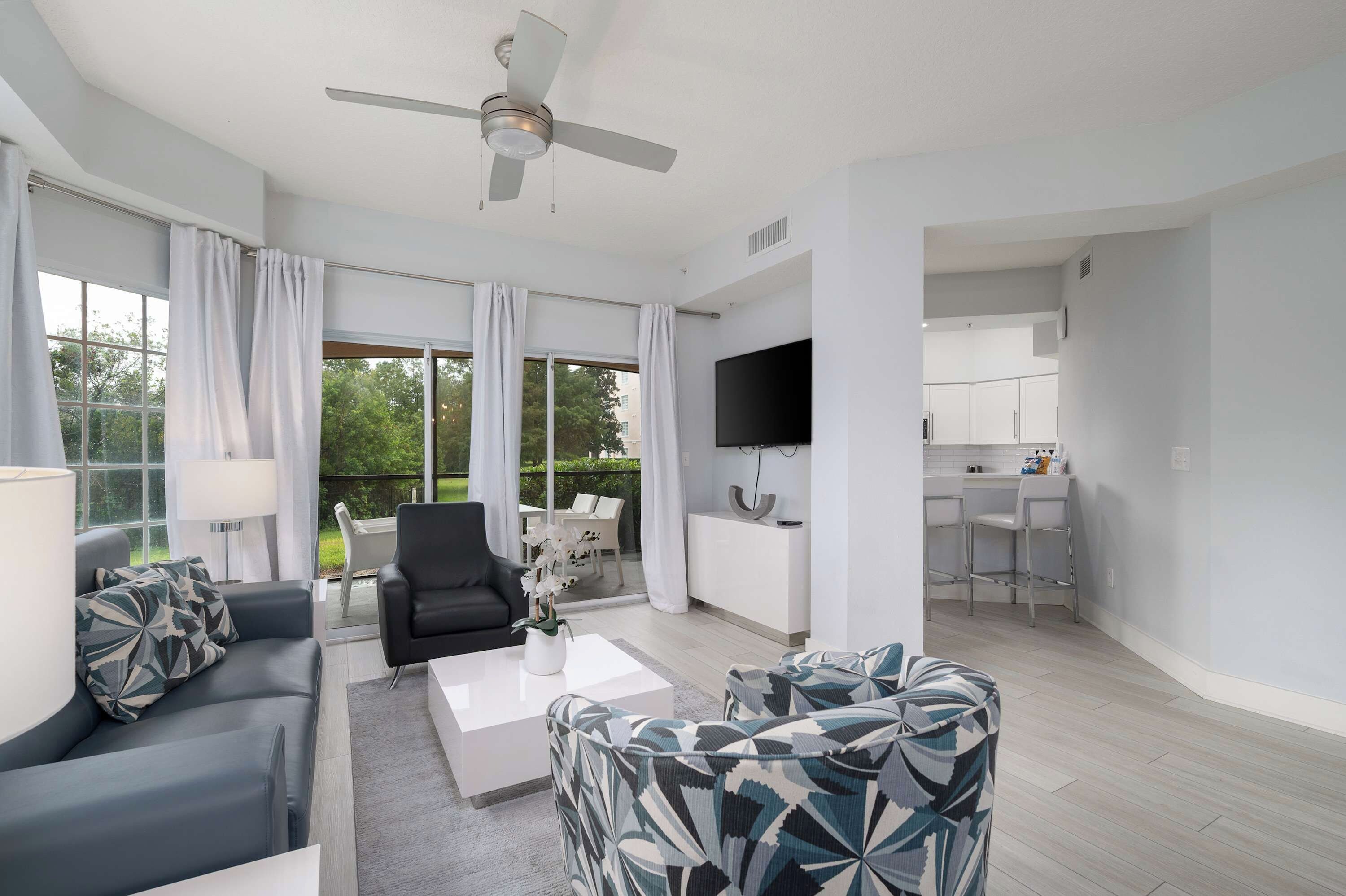 A modern living room with a sofa, coffee table, and patterned armchairs. A ceiling fan and a flat-screen TV are mounted on the wall.