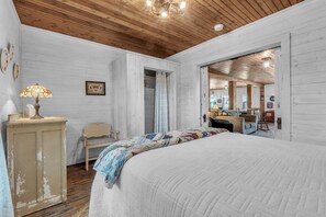 Ein Schlafzimmer mit Holzboden, einem Bett mit Quiltdecke, einem Nachttisch, einem Sessel und einer Tür zu einem anderen Raum.