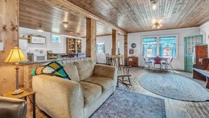 Ein gemütliches Wohnzimmer mit einer Couch, einem Couchtisch und einem Teppich. Es gibt einen Essbereich mit Tisch und Stühlen. Das Zimmer verfügt über Holzbalken an der Decke und ein Fenster mit Vorhängen.