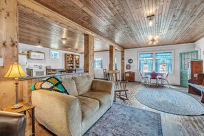 Ein gemütliches Wohnzimmer mit einer Couch, einem Couchtisch und einem Teppich. Es gibt einen Essbereich mit Tisch und Stühlen. Das Zimmer verfügt über Holzbalken an der Decke und ein Fenster mit Vorhängen.