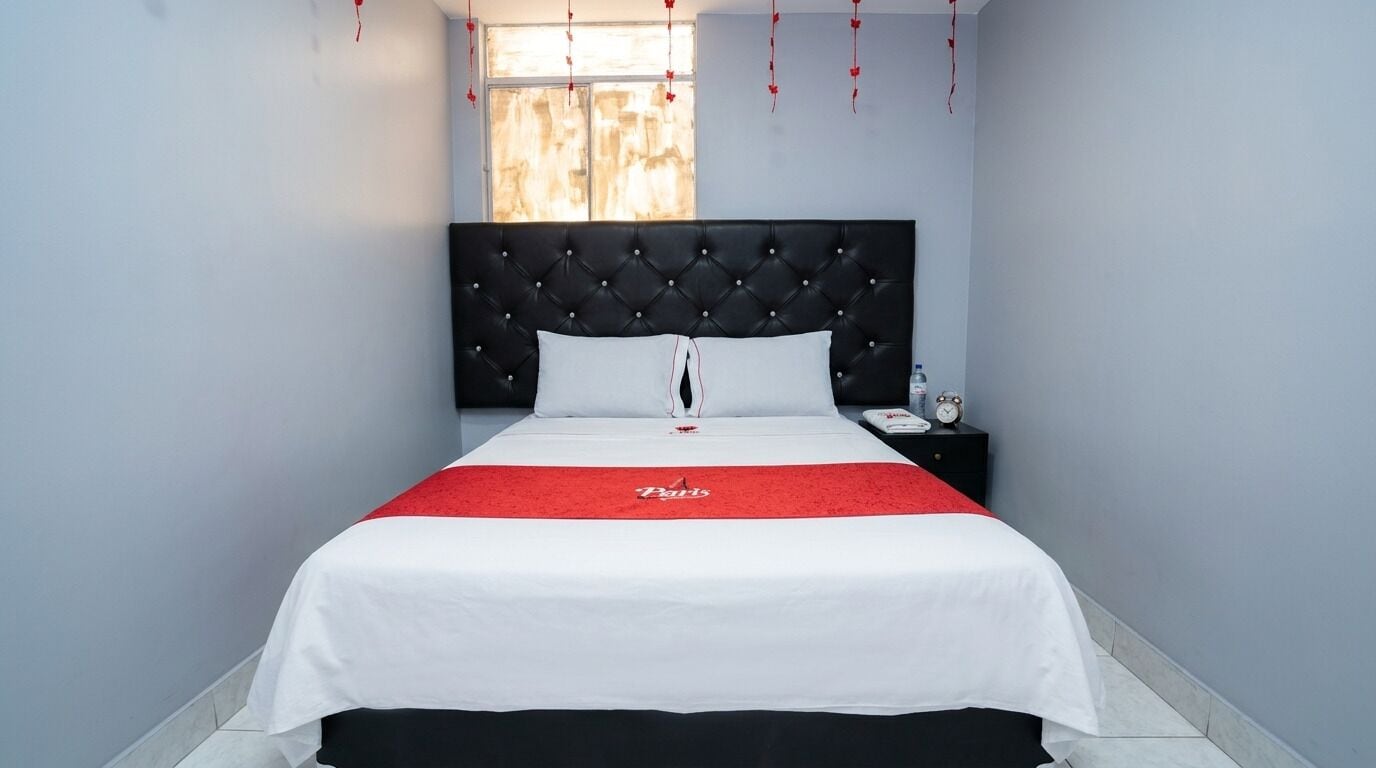 A hotel room with a bed, a nightstand, a window, and a red bedspread.