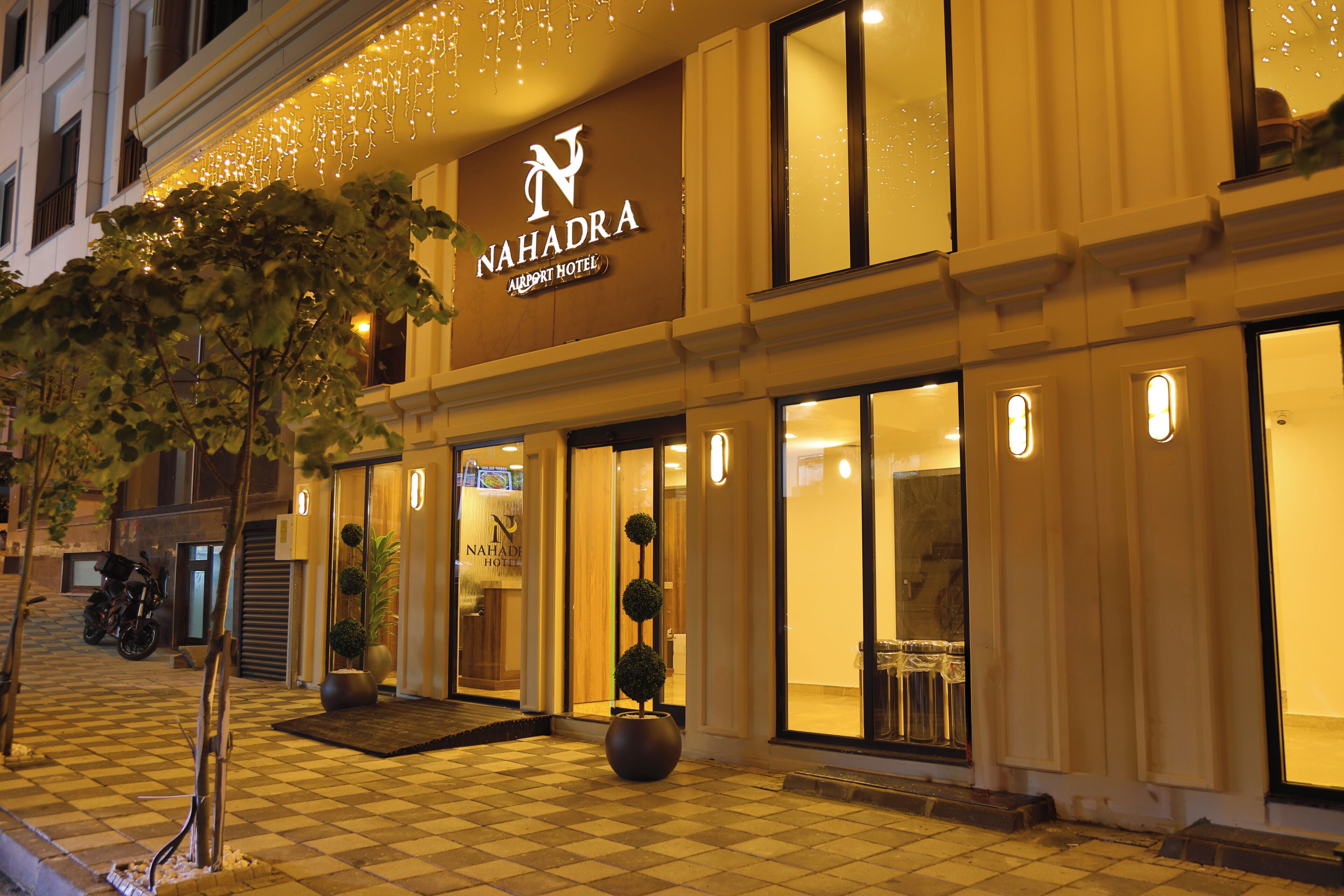 A hotel entrance with a checkered floor, potted plants, and a sign reading 'NAHADRA AIRPORT HOTEL'.