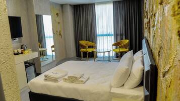 Quarto de hotel com cama, duas cadeiras, escrivaninha, televisão de tela plana e uma janela grande com cortinas.