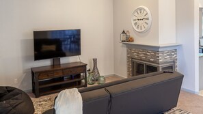 A living room with a flat-screen TV on a wooden stand, a fireplace with a clock above it, and a dark-colored sofa. - Corporate executive condo! Newly furnished in quiet gated community. (Arlington)