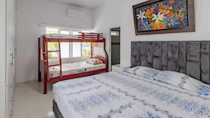 A bedroom with a bunk bed, a large bed with patterned bedding, a grey upholstered headboard, and a colorful painting on the wall.