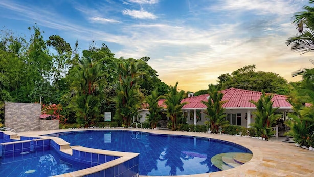 A swimming pool with a curved edge, surrounded by lush greenery and a building with a red roof.