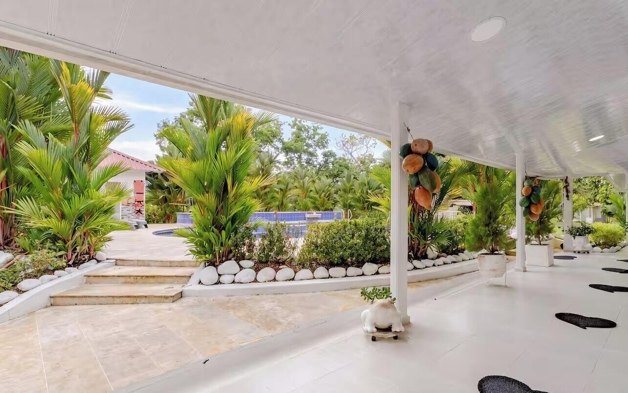 An indoor covered area with a view of a swimming pool and palm trees, featuring a white floor and steps leading to the pool.