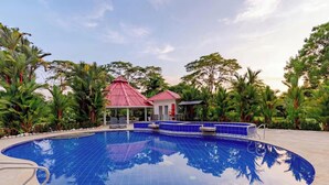 A swimming pool with a red-roofed pavilion, surrounded by palm trees and a clear sky.
