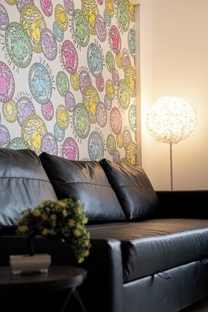 Ein modernes Wohnzimmer mit einem schwarzen Ledersofa, einem kleinen Beistelltisch mit Pflanze und einer Wand mit bunt gemustertem Wandpapier.