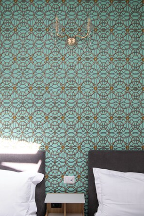 Ein Zimmer mit gemustertem Tapeten, zwei Betten, einem kleinen Nachttisch und einer Wandleuchte.