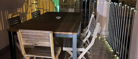 Un patio al aire libre con una mesa y sillas de madera, una barandilla de metal y un edificio de ladrillo al fondo.