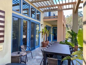 A covered outdoor patio with a long table and chairs, large windows, and potted plants. - River Villa with pool and spa ~ Best Swimming Hole (Three Rivers)