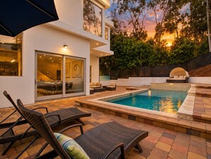 A modern outdoor pool area with lounge chairs, a glass door entrance, and a view of the sunset. - ৎ୭Luxury Hollywood Hills | 5BR + Poolৎ୭ (Los Angeles)