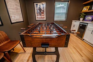 A foosball table in a room with a wooden floor, a poster on the wall, and a window with blinds. - North Hills Raleigh Retreat (Raleigh)
