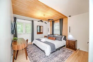 Une chambre moderne avec un plafond en bois, un grand lit avec une literie grise, une petite table avec une plante et un tapis à motifs.