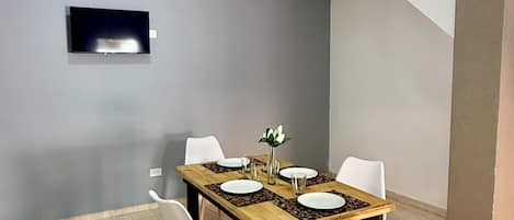 Un comedor con una mesa dispuesta para cuatro personas, sillas blancas, un televisor de pantalla plana montado en la pared y una escalera que conduce a otro nivel.