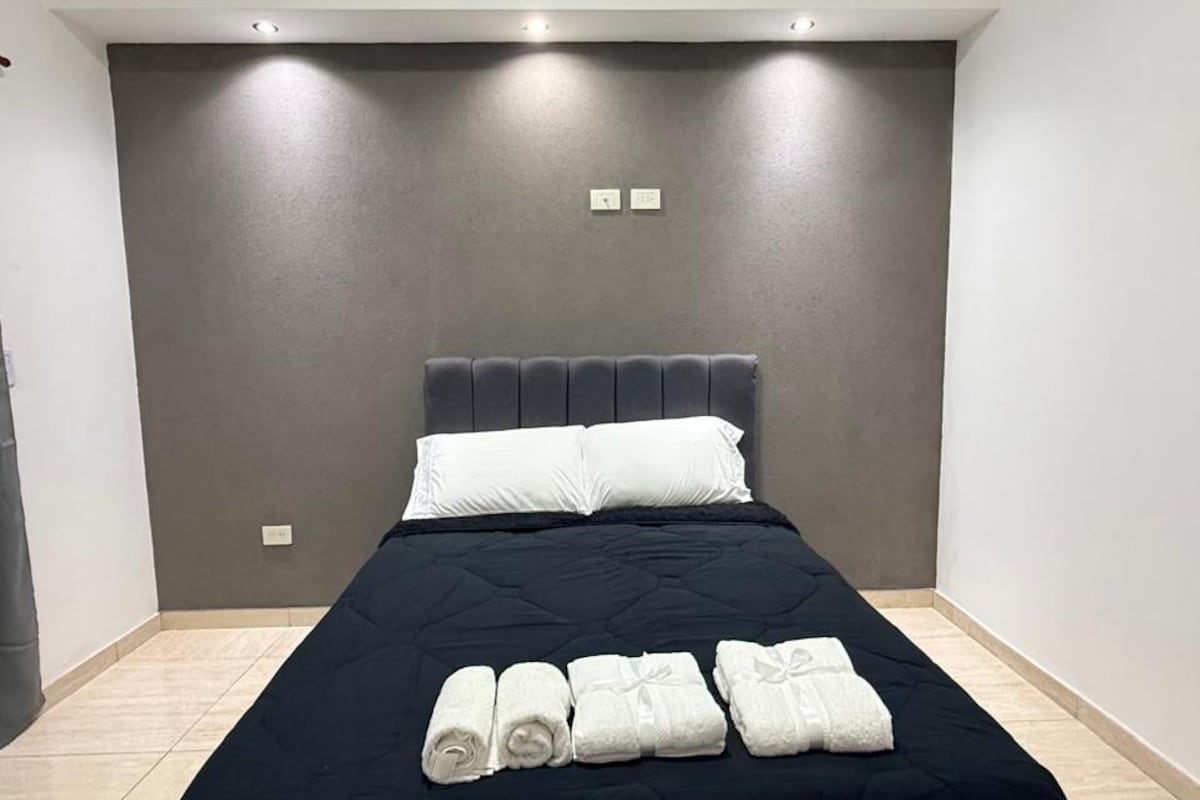 A neatly made bed with a dark bedspread and white pillows, a wall-mounted light fixture above, and a plain wall with a small electrical outlet.