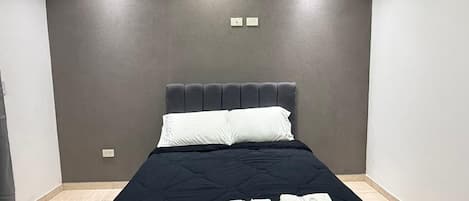 A neatly made bed with a dark bedspread and white pillows, a wall-mounted light fixture above, and a plain wall with a small electrical outlet.
