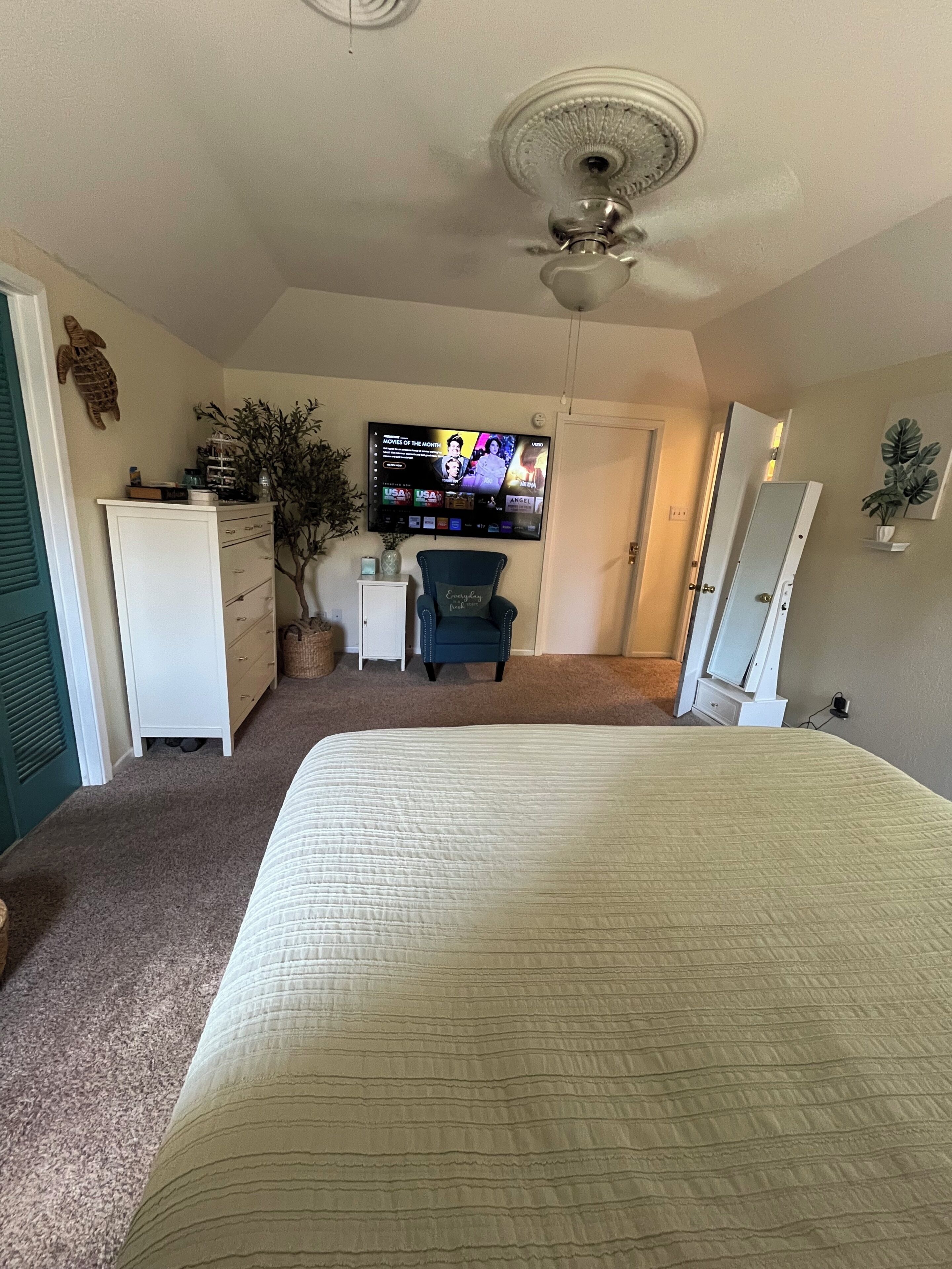 Une chambre à coucher comprenant un lit, une commode, une chaise, un téléviseur fixé au mur et un ventilateur de plafond.
