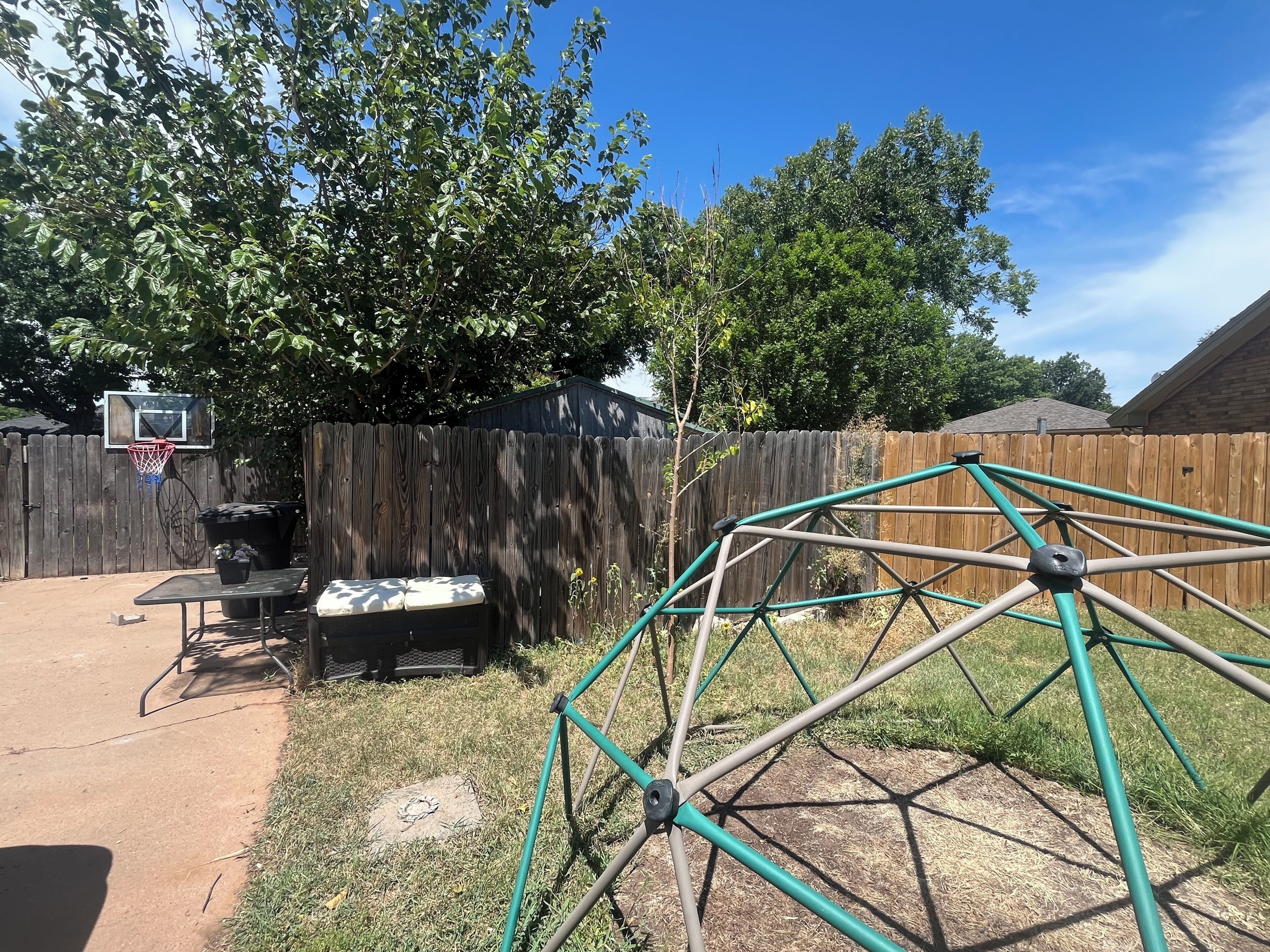A backyard with a basketball hoop, a small table with cushions, and a green metal structure.