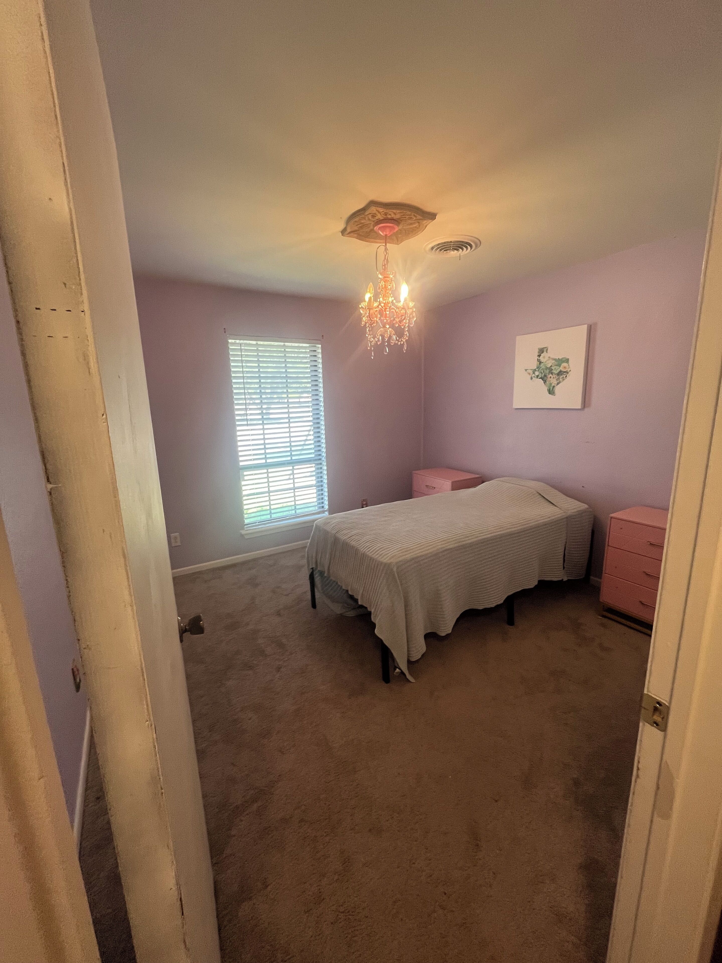 A bedroom with a bed, a dresser, a chandelier, a window with blinds, and a wall-mounted picture.