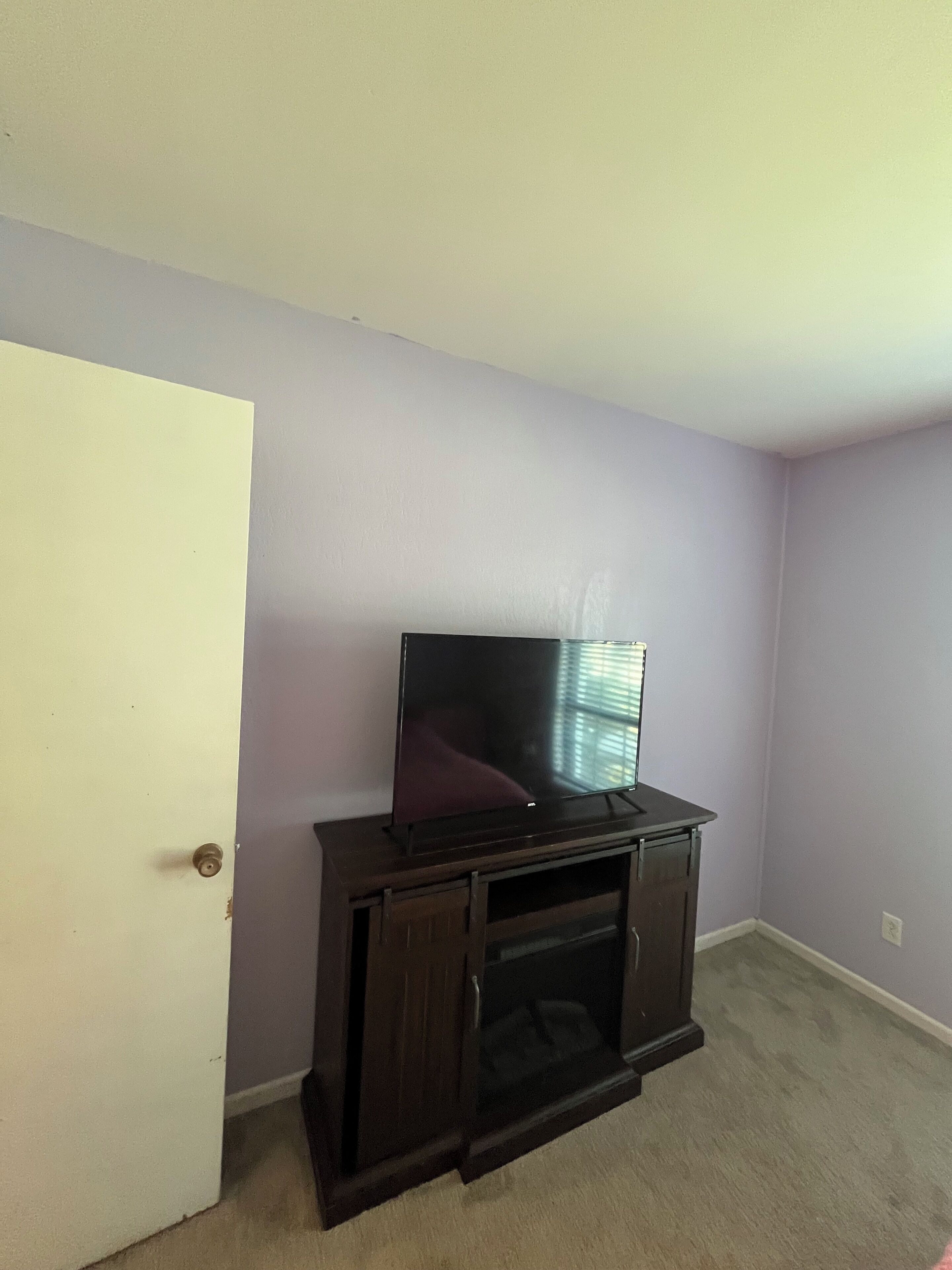 Una habitación con una televisión sobre un soporte de madera, una puerta blanca y paredes de color lila claro.