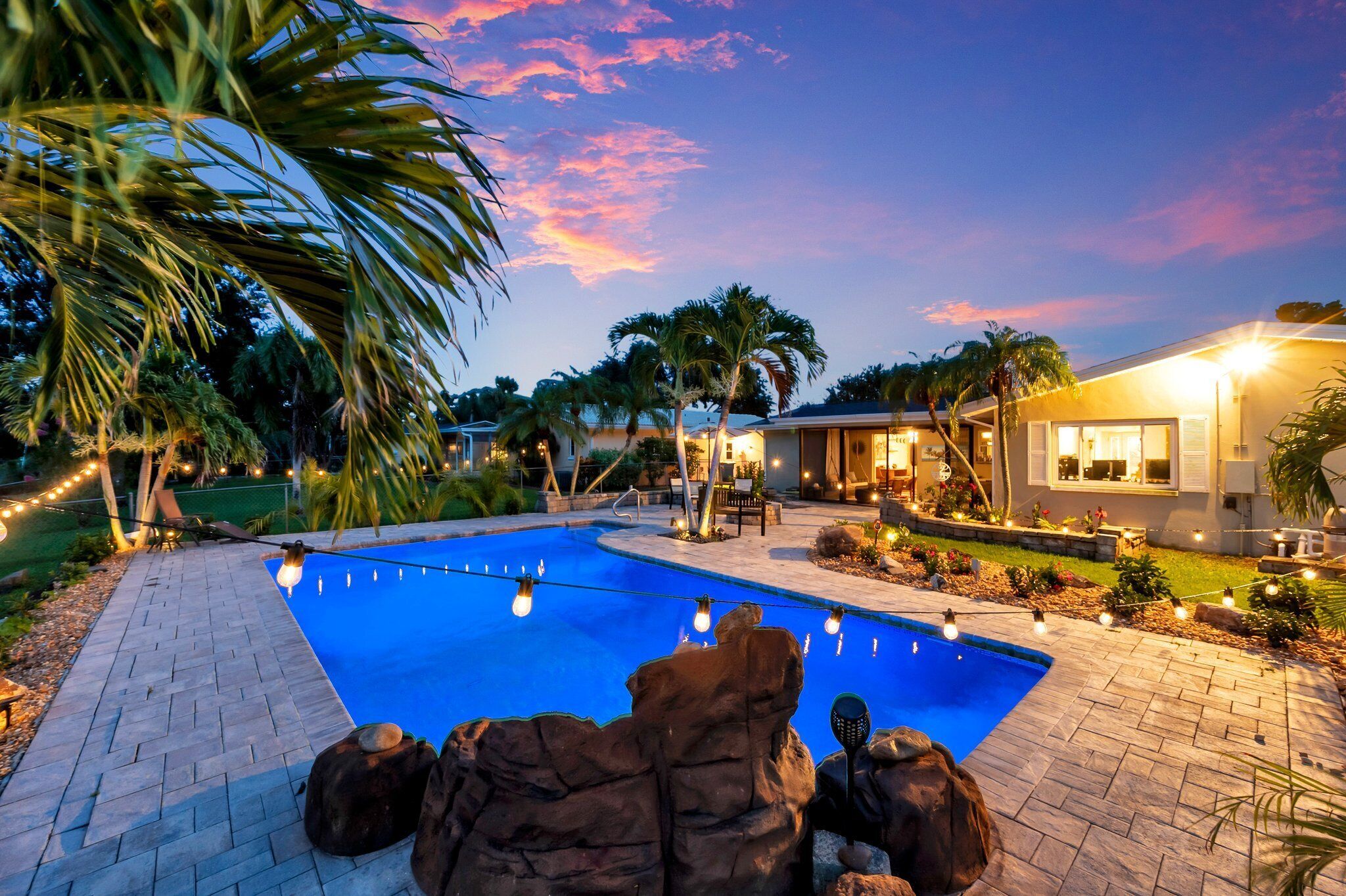 A swimming pool with a paved deck, surrounded by palm trees and a house with a visible patio area.