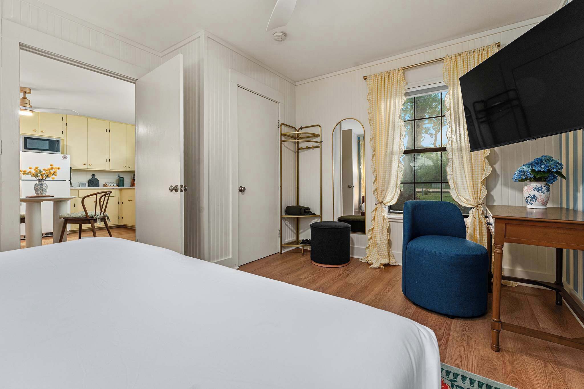 A hotel room with a bed, a blue armchair, a small table, a mirror, and a view of the kitchen.