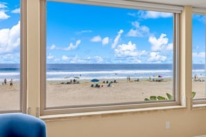 Ein Zimmer mit großem Fenster und Blick auf einen Strand mit Menschen und Sonnenschirmen.