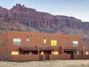 A modern building with a flat roof and multiple windows, situated in a desert landscape with a mountain in the background.