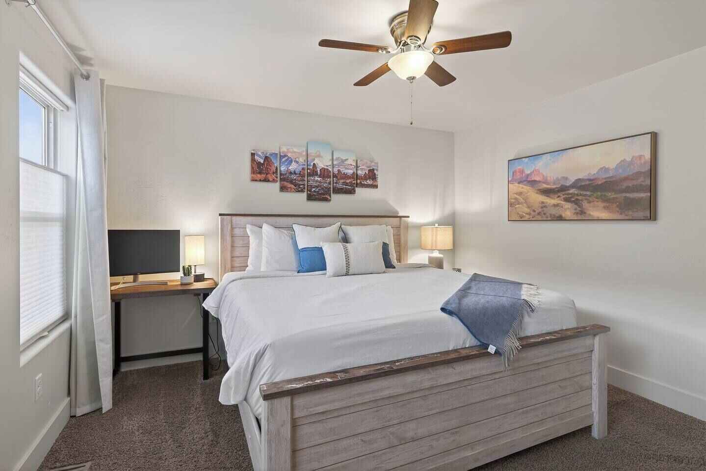 A bedroom with a bed, a desk, a chair, a ceiling fan, and artwork on the walls.