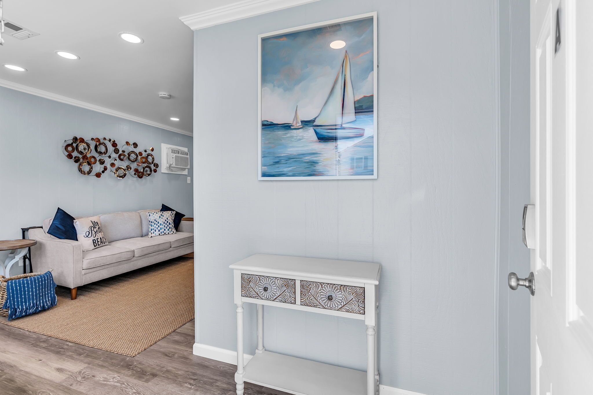 A living room with a light blue wall, a white sofa, a patterned console table, and a painting of sailboats.