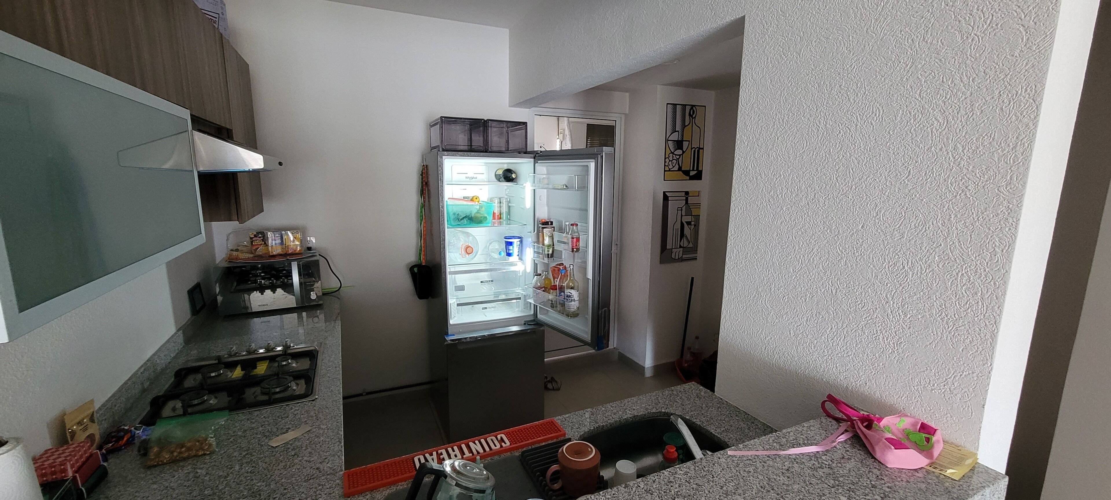 Een keuken met een koelkast, een magnetron en een spoelbak. Er staan verschillende spullen op het aanrecht, waaronder een fles, een beker en een zak.
