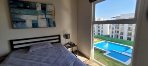 Une chambre avec une grande fenêtre donnant sur une piscine et des immeubles d’habitation.