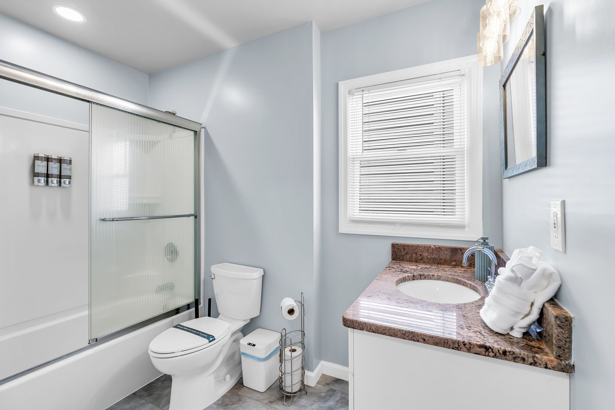 Une salle de bain moderne avec une douche à parois de verre, des toilettes, un lavabo doté d’un plan de travail en marbre et une fenêtre avec des stores.