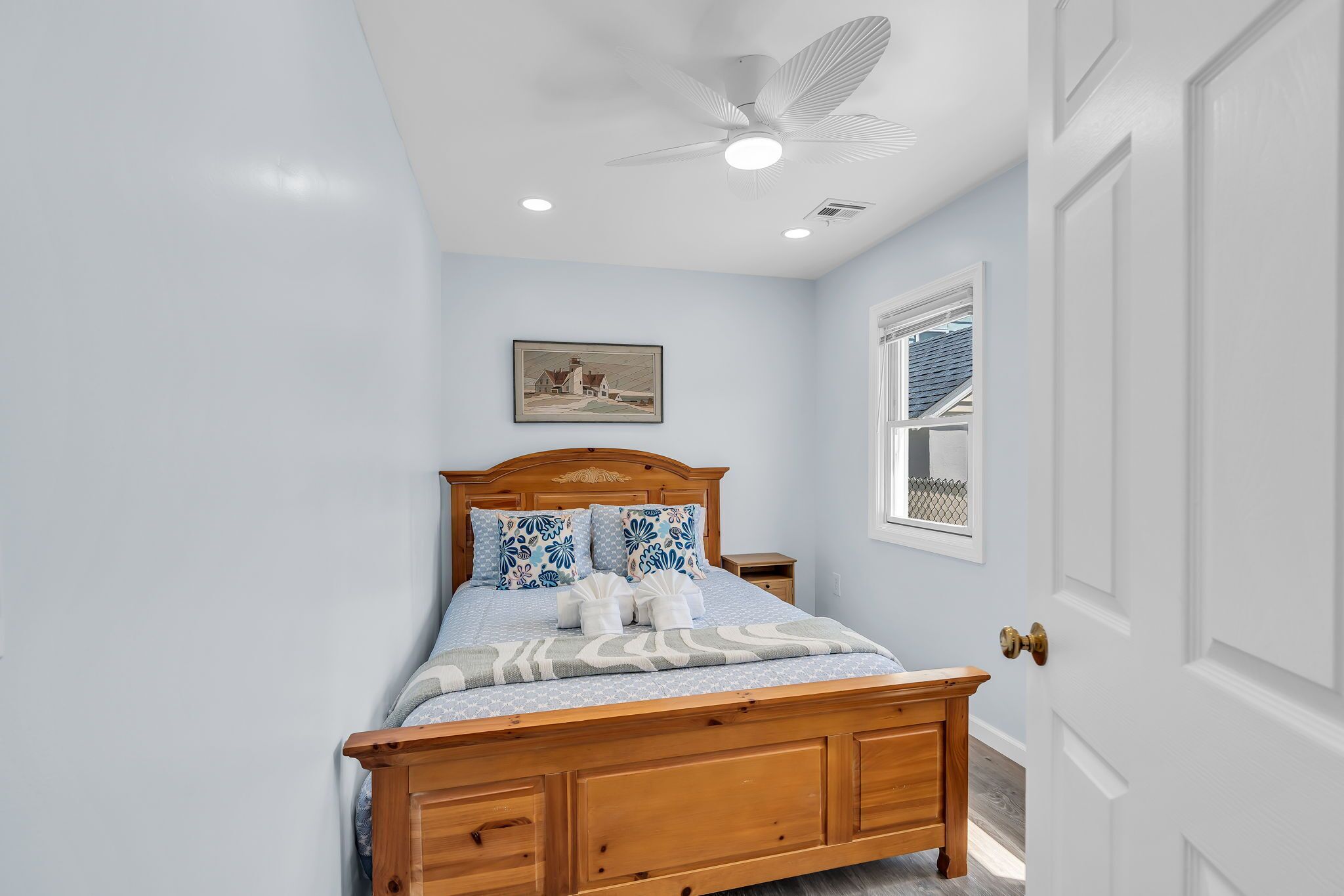 A bedroom with a wooden bed, a ceiling fan, recessed lighting, a window with a view, and a framed picture on the wall.