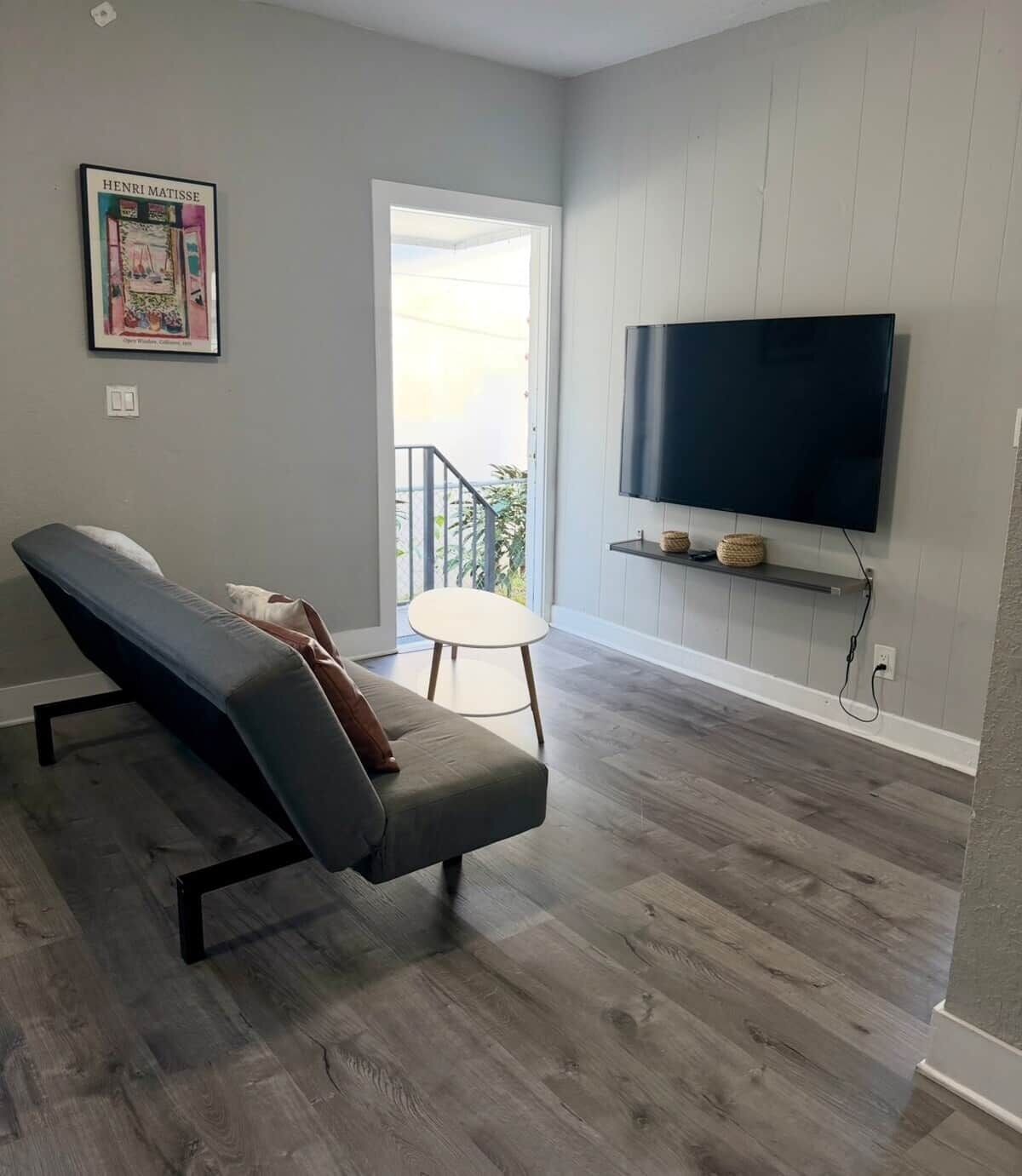 Ein modernes Wohnzimmer mit einem grauen Sofa, einem flachbildfernseher an der Wand und Holzboden.