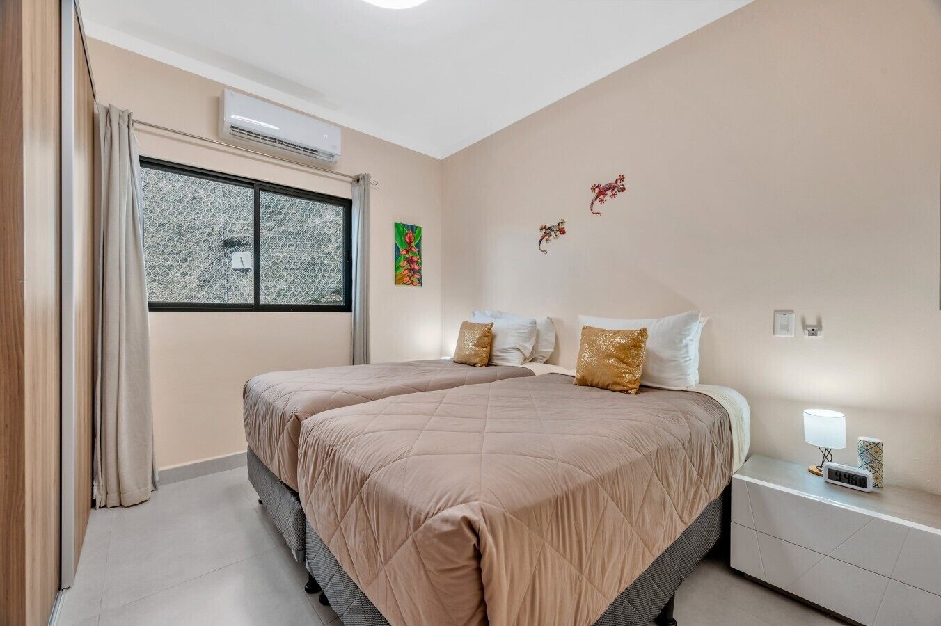 A bedroom with two beds, a window with patterned curtains, an air conditioning unit, and a bedside table with a lamp and a clock.