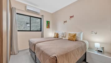 A bedroom with two beds, a window with patterned curtains, an air conditioning unit, and a bedside table with a lamp and a clock.
