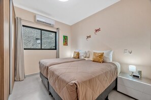 A bedroom with two beds, a window with patterned curtains, an air conditioning unit, and a bedside table with a lamp and a clock. - Villa Ladera Laurel 10 | Luxe Hillside Retreat with Epic Views (Potrero)