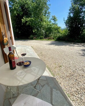 Una mesa con una botella de vino y copas, un bol de aperitivos y vistas a un camino de grava que lleva a un jardín con árboles.